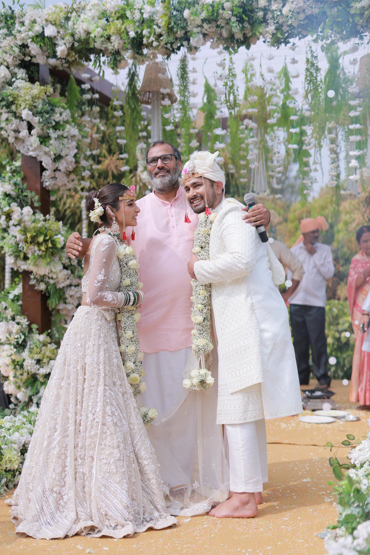 Hemant Acharya with his arms around a bride and groom under a white floral mandap, microphone still in hand