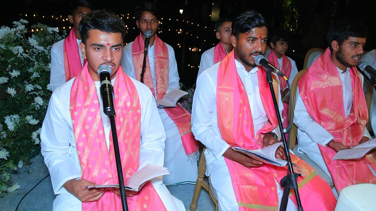 Pandits chanting from ritual books into microphones at an evening ceremony lit by strings of lights