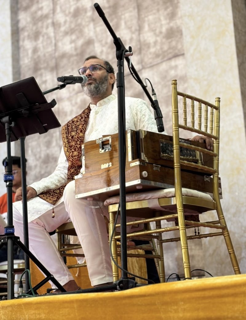 Hemant Acharya in a white chikankari kurta and maroon jacket, singing at the harmonium